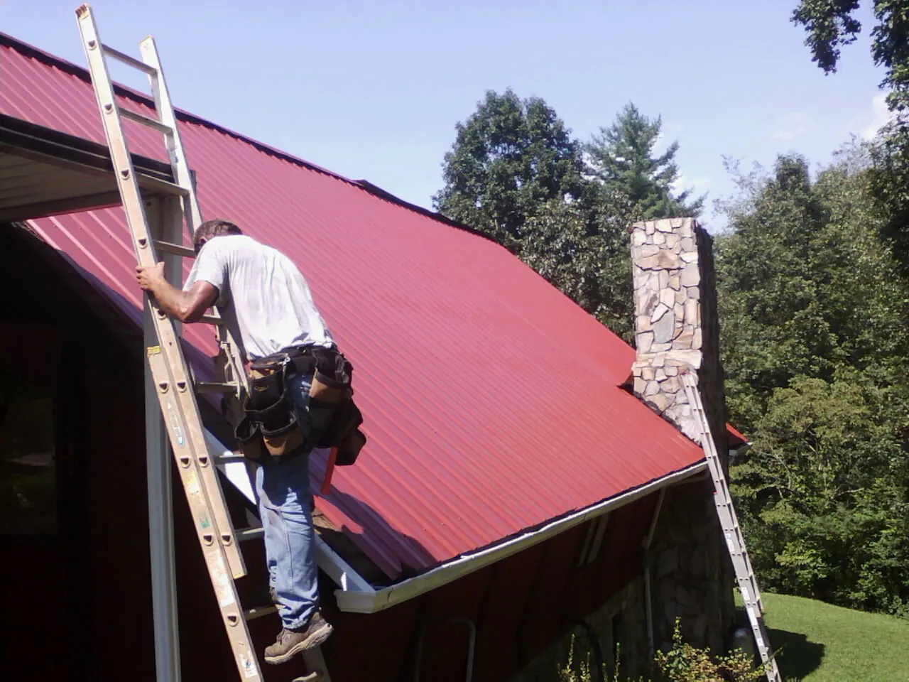 Roofer on a ladder beside a red metal roof and stone chimney — Soffit & Fascia Repair by Roofing Brothers in Wiota